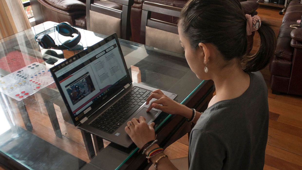 Photo of a woman seen from  behind working on a laptop at a glass table.