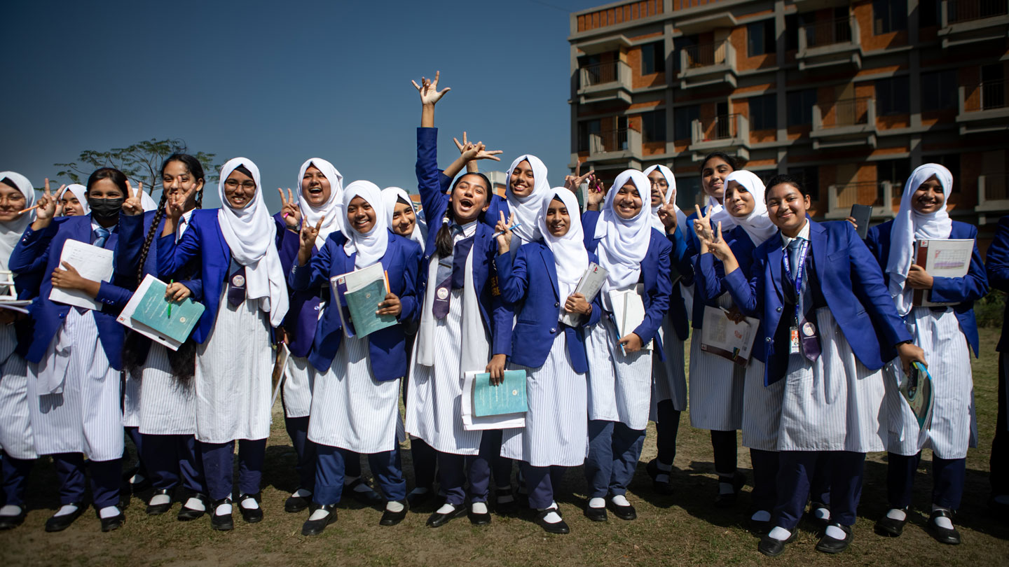 Photo of บังกลาเทศi schoolgirls in uniform smiling and holding textbooks outside their school building.