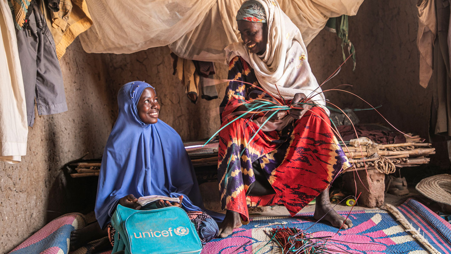 Photo of a girl in a blue outfit with a UNICEF bag sitting indoors with a woman in colorful traditional clothing.