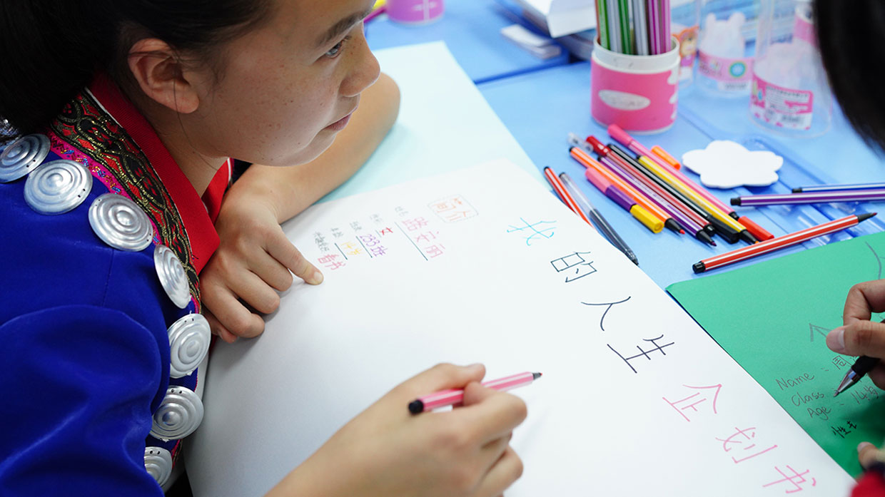 Photo of a girl in traditional clothing writing with colored pens on a sheet of paper with Chinese characters.