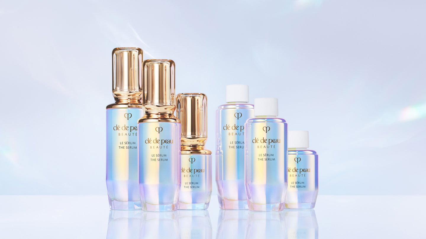 Photo of multiple bottles of Cl&eacute; de Peau Beaut&eacute; THE S&Eacute;RUM in various sizes, featuring iridescent packaging with gold or white caps.