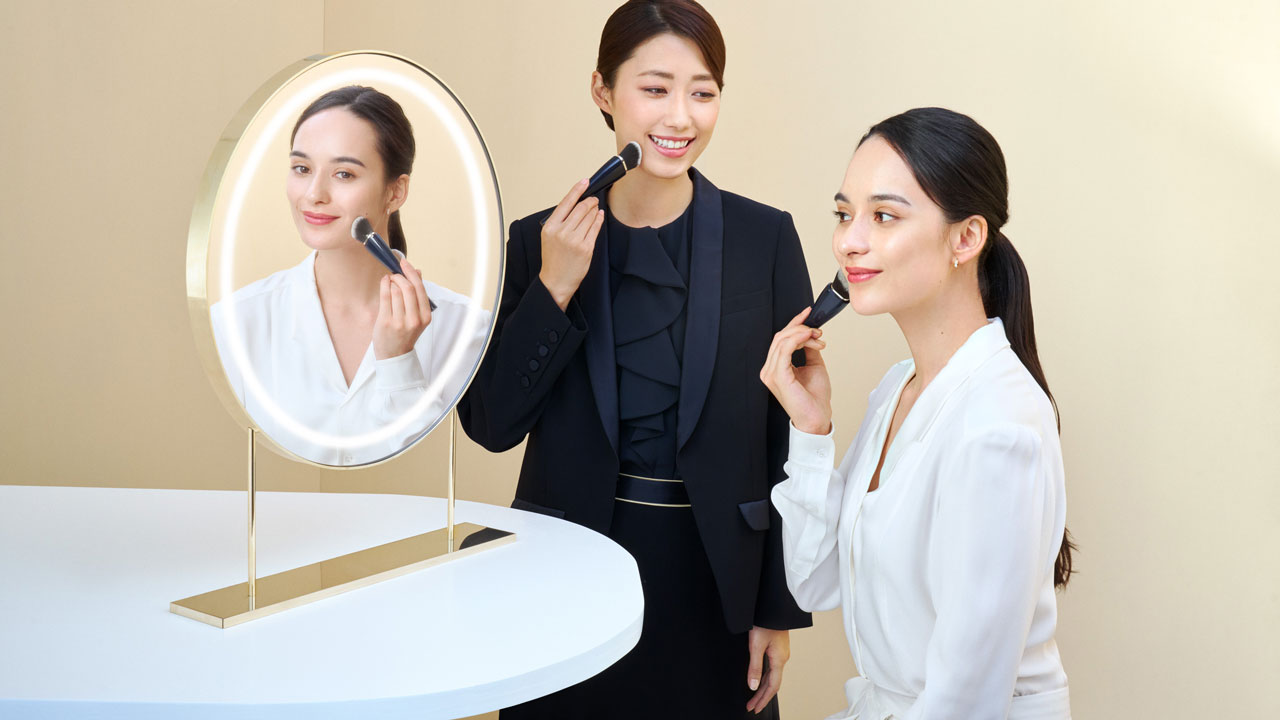 Photo of a personal beauty specialist providing makeup guidance to a woman in front of a round mirror with built-in lighting.