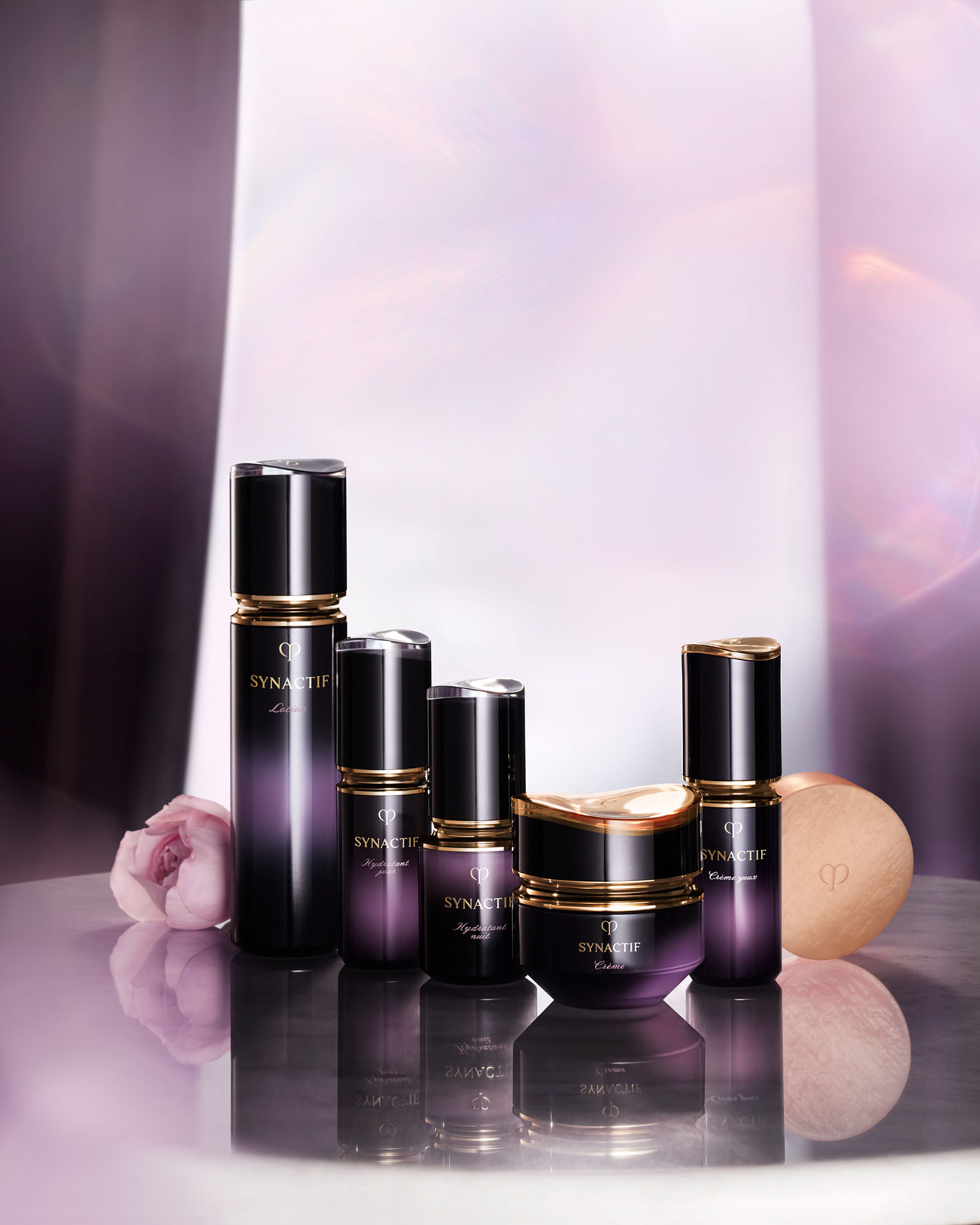 Photo of the Cl&eacute; de Peau Beaut&eacute; Synactif skincare lineup, arranged from left to right as: Lotion, Daytime Moisturizer, Nighttime Moisturizer, Cream, Eye Cream, and Soap &mdash; all in gradient black-to-purple packaging with gold accents.