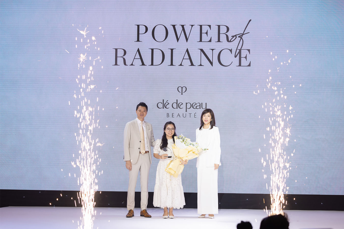Photo of three people on stage, centered around a person holding a bouquet, with fireworks effects on both sides.