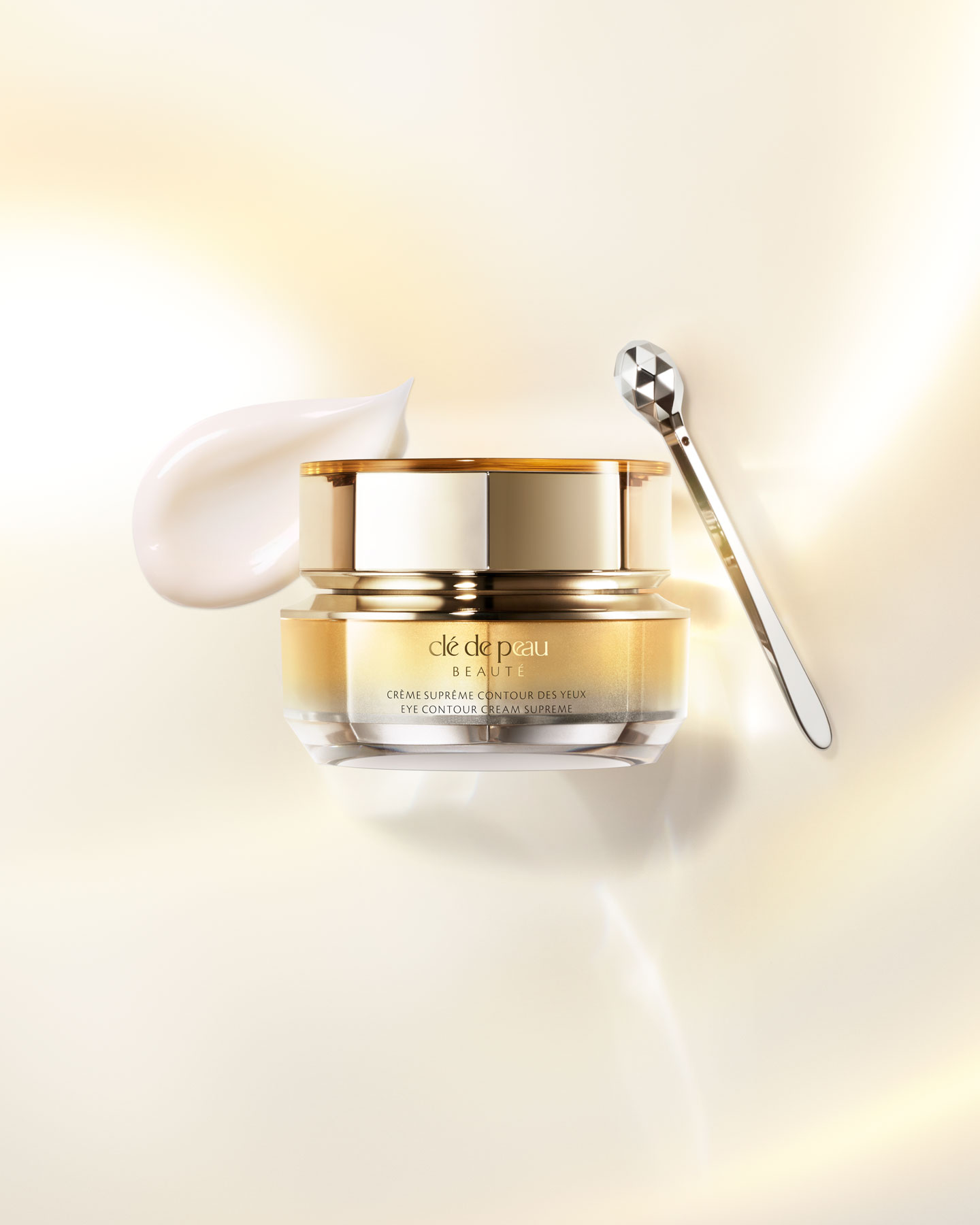 Photo of an EYE CONTOUR CREAM SUPREME jar with a gold-to-silver gradient and an accompanying spatula.