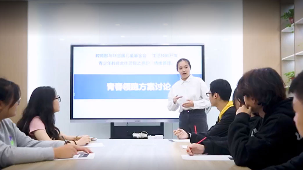 Photo of a woman in presenting a conference room using a screen, while several participants listen attentively arround the table.