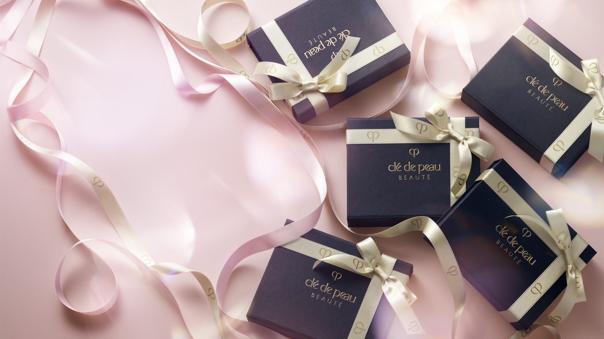 Wallpaper of Cl&eacute; de Peau Beaut&eacute; dark blue gift boxes arranged on a soft pink background, with champagne gold and pink ribbons featuring the brand logo elegantly intertwined.