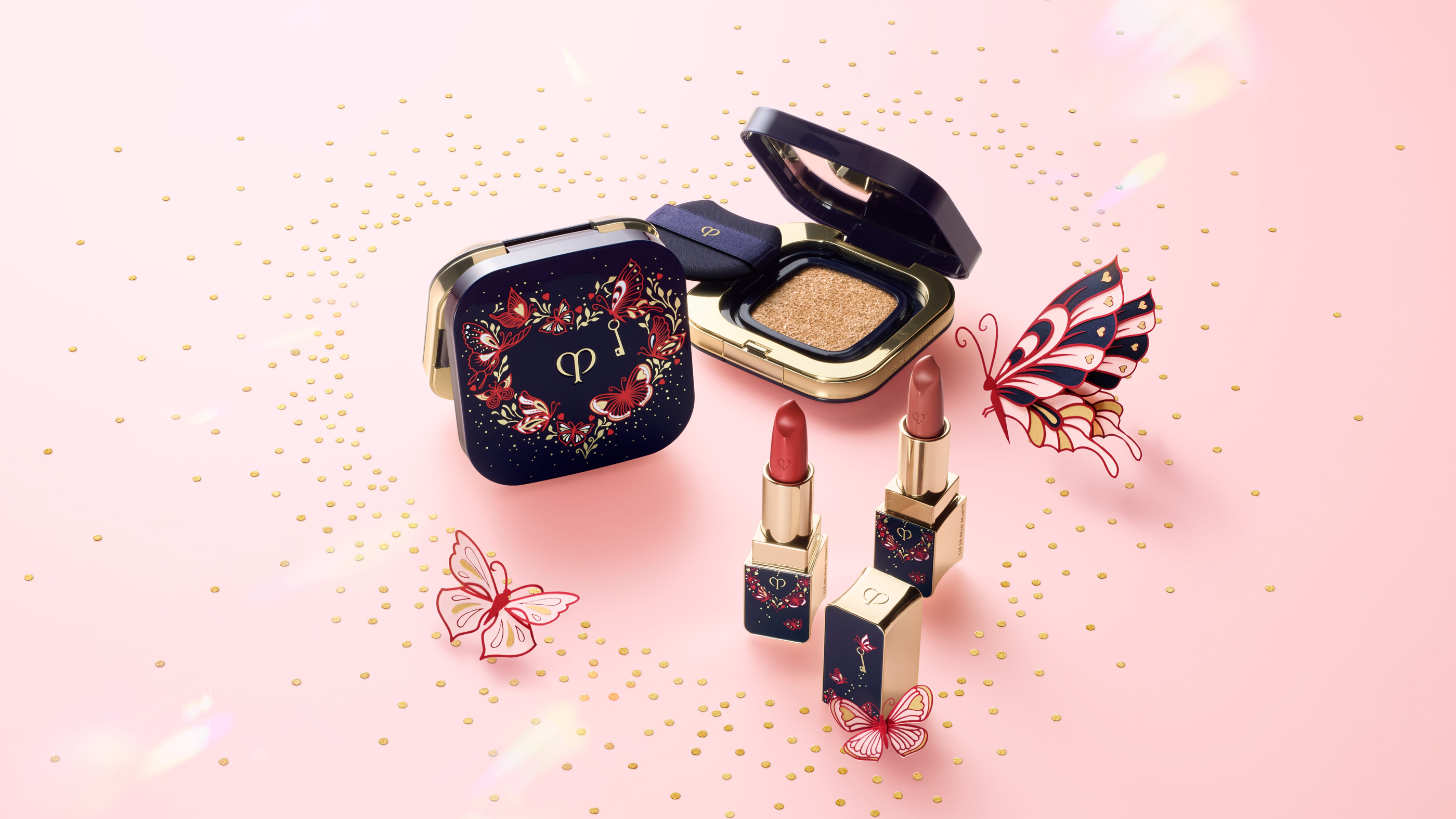 Photo of a compact and lipsticks from the 26SS limited-edition Butterfly Kisses collection are displayed on a pink background sprinkled with golden dots. The blue-black packaging features a gold heart adorned with delicate red butterfly motifs and a small gold key.