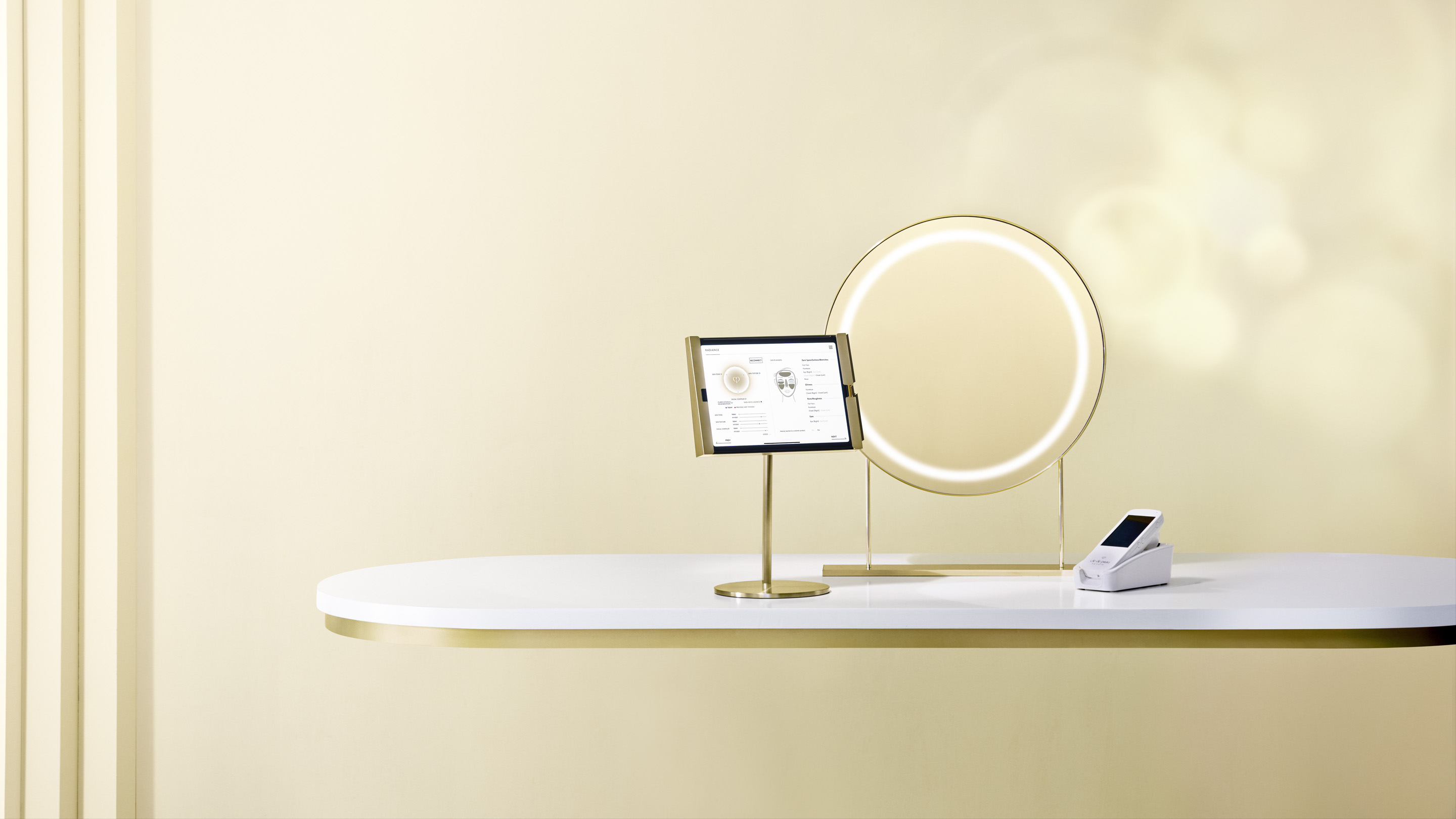 Photo of the Cl&eacute; de Peau Beaut&eacute; Smart Radiance Mirror setup, showing a gold-framed ring mirror, an iPad connected to a Handy Skin Sensor (HSS), and a white HSS device placed on a clean display table.