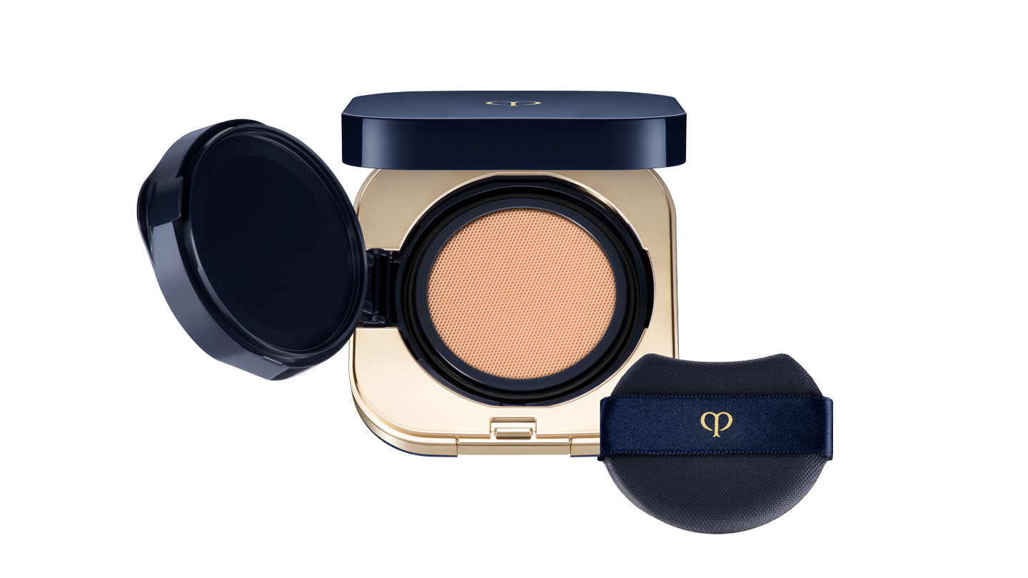 Photo of RADIANT CUSHION FOUNDATION NATURAL. The compact is opened to reveal a cushion foundation with a smooth surface, housed in a glossy gold and deep navy case. A black puff with the brand logo is included.