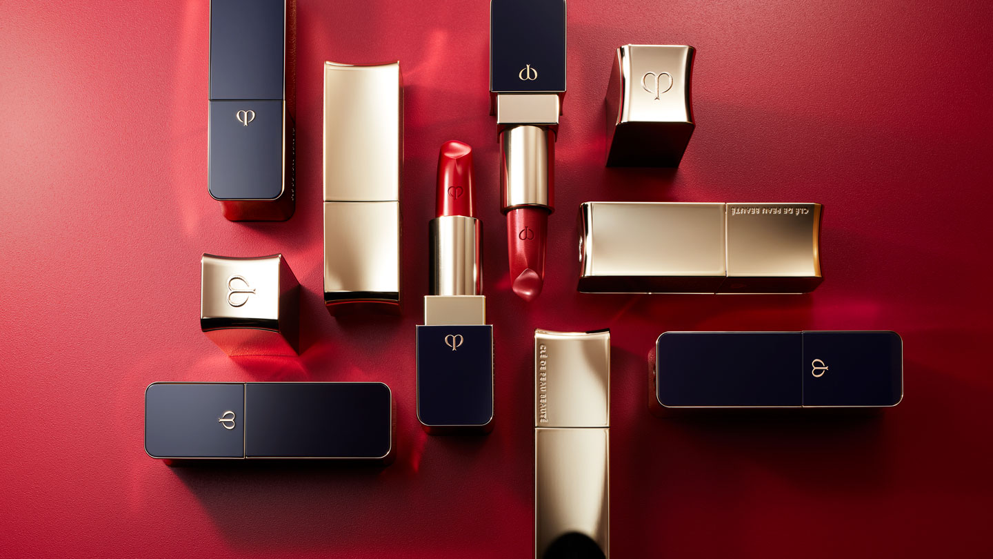 Photo of a vivid red lipstick with its cap removed at the center, surrounded by other lipsticks with their caps on, arranged elegantly on a deep red background.