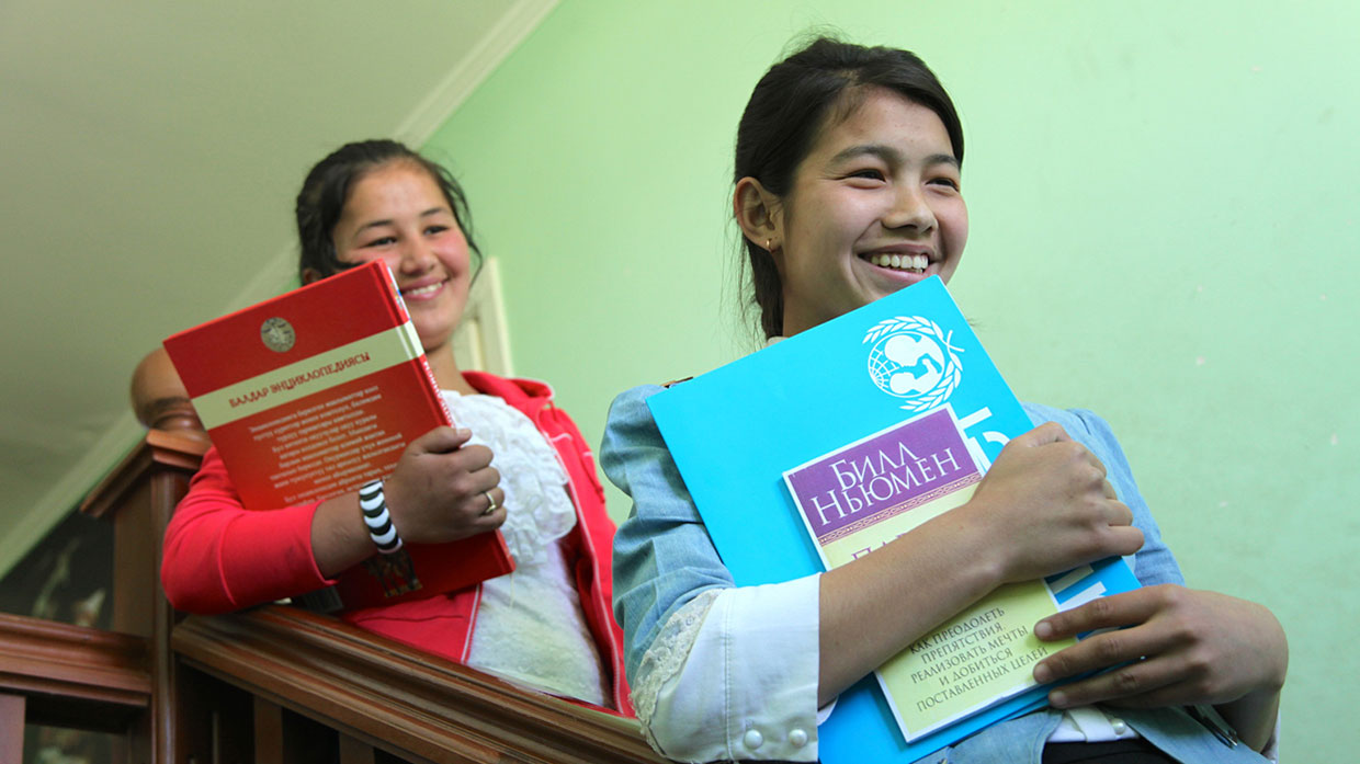 Photo of two women smiling while standing on a staircase, holding educational materials featuring the UNICEF logo.