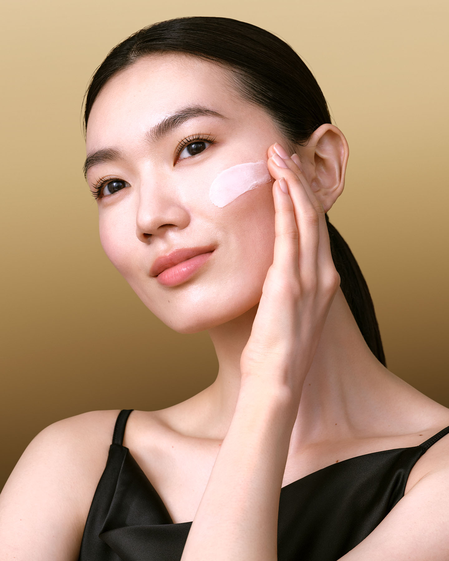 Photo of the BRIGHTENING SERUM SUPREME. The prismatic glass bottle with a geometric gold cap stands on a minimalist pedestal against softly curved grey and white panels. To the right, two drops of the creamy serum texture are shown suspended in mid-air.