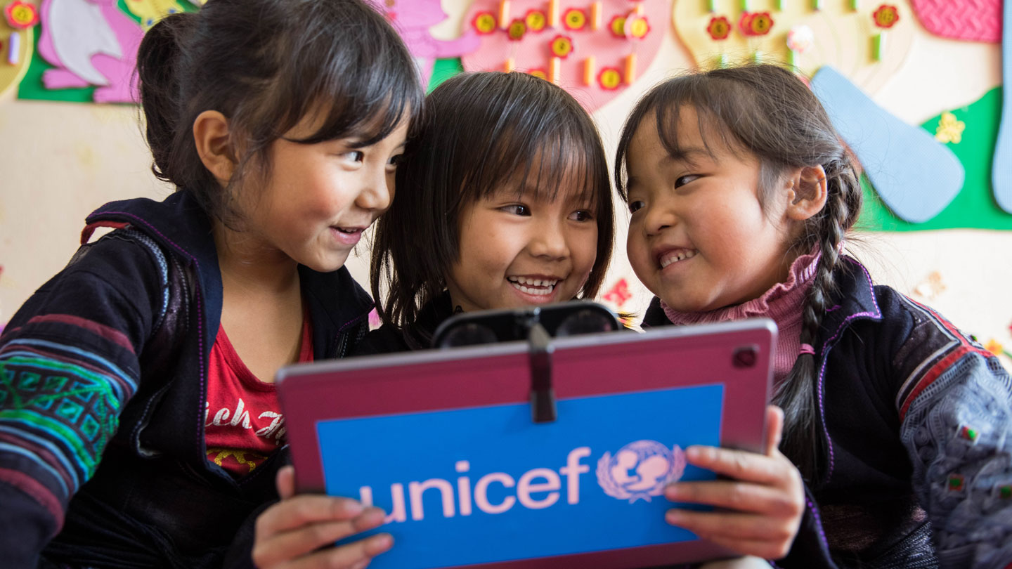 Photo of three children smiling as they look at a tablet with the UNICEF logo.