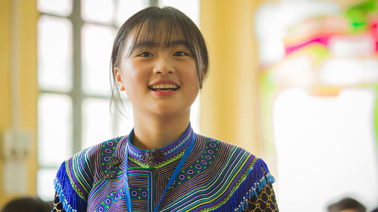 Photo of a woman wearing traditional ethnic attire, smiling in a classroom-like setting.
