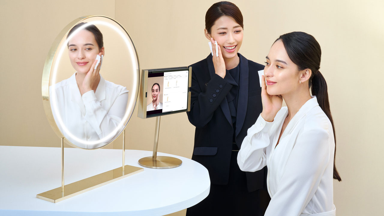 Photo of a personal beauty specialist guiding a woman on skincare techniques in front of a round mirror with built-in lighting.