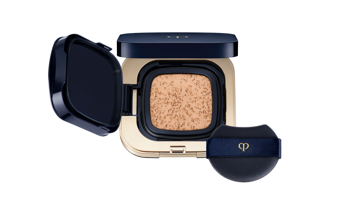 Photo of RADIANT CUSHION FOUNDATION DEWY. The compact is opened to reveal a dewy-textured cushion foundation housed in a glossy gold and deep navy case. A black puff with the brand logo is included.