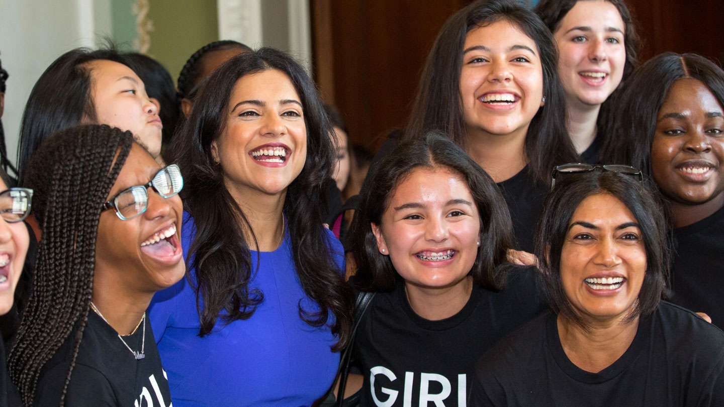 Photo of Reshma Saujani laughing with a group of diverse young girls, all smiling and waring black T-shirts with the word "GIRL" printed in white.