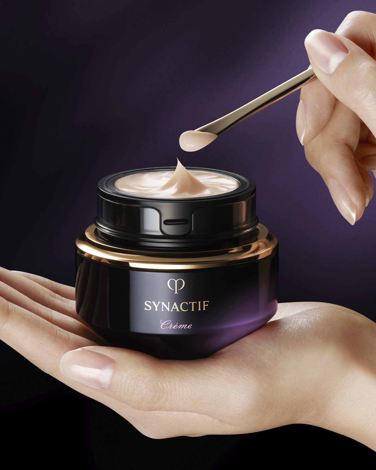 Photo shows a hand holding a jar of SYNCACTIF CREAM with a black-to-purple gradient and gold accents. Another hand is using a gold spatula to scoop out some of the cream. 